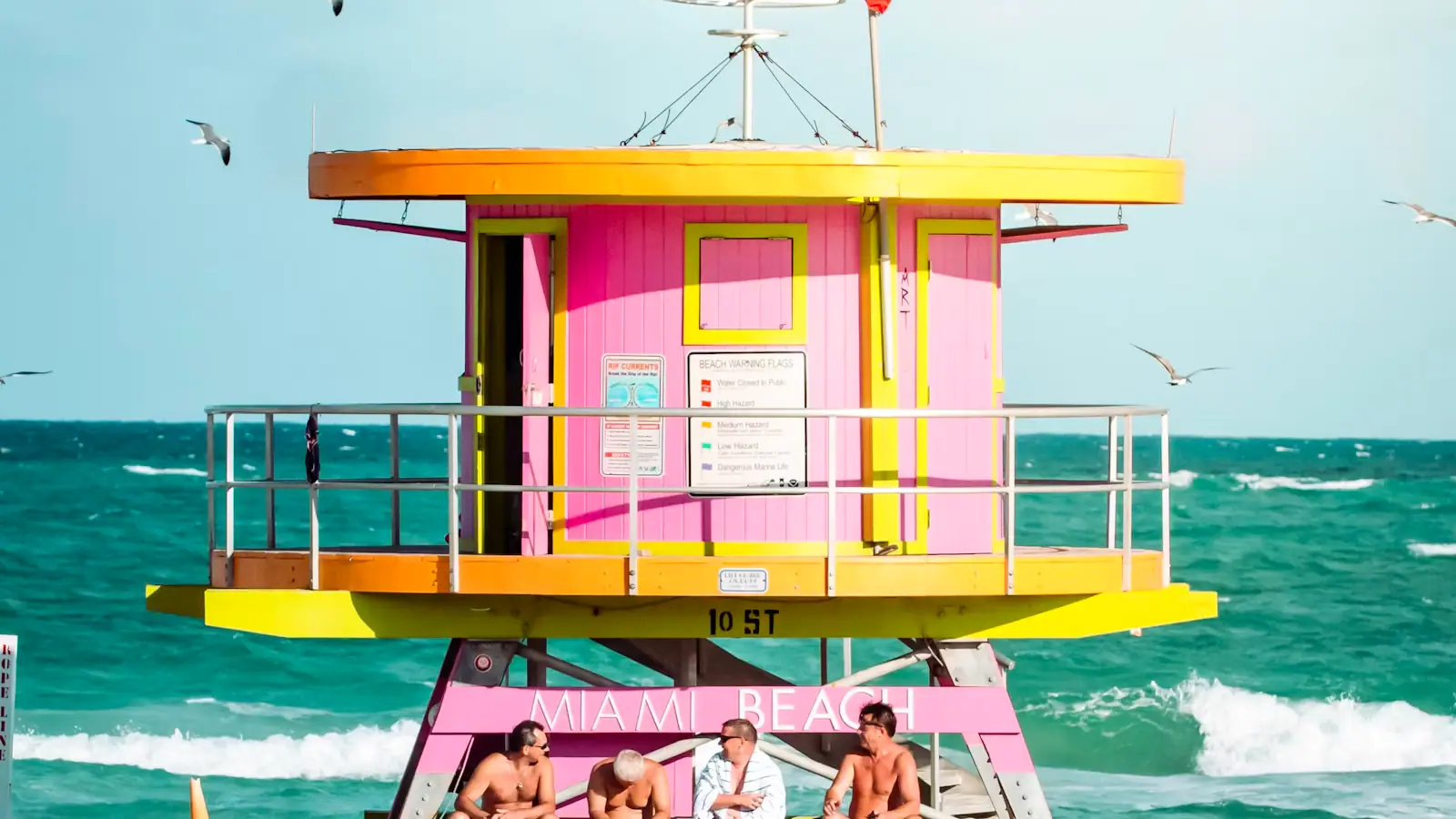 A pink and yellow lifeguard tower on South Beach with people sitting in the shade beneath it
